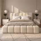 A modern upholstered bed with a chunky segmented headboard and matching footboard in a warm cream bouclé fabric, styled in a minimalist beige bedroom with white pillows, neutral bedding, matching nightstands, and hanging pendant lights.