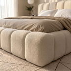 A close-up of a modern bed with a chunky, tufted upholstered frame in a warm cream bouclé fabric, dressed in neutral beige bedding and a textured throw blanket, set in a softly lit bedroom.