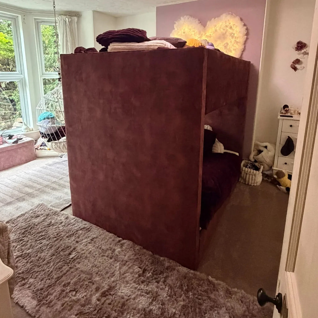 A dark reddish-brown upholstered loft bed or bunk bed frame in a child's bedroom, with bedding on top, a window, hanging chair, rug, and decorative wall art visible.
